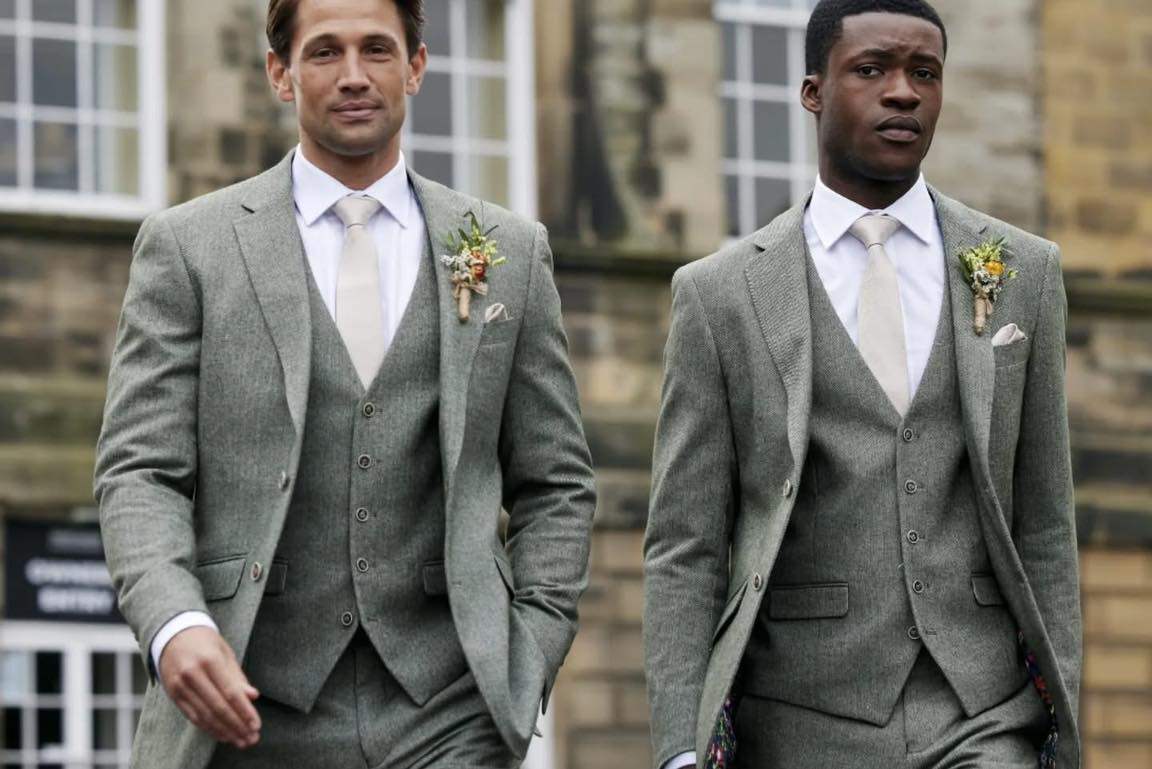 Two grooms in sage tweed three-piece suits in front of the Alnwick sandstone, photographed by Bell and Sons