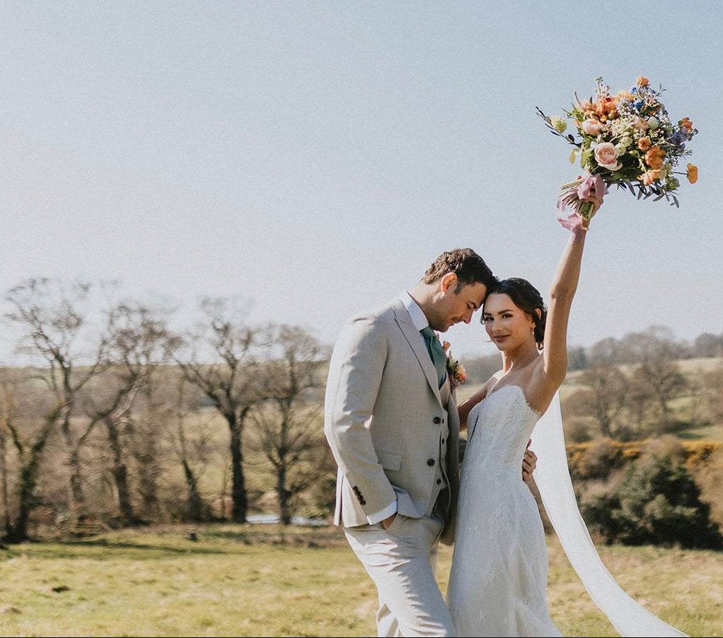 A groom and bride in front of an open Northumberland field, the groom in a stone three-piece suit fitted from Bell and Sons